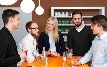 Fünf junge Menschen stehen an einem Holztisch und unterhalten sich.